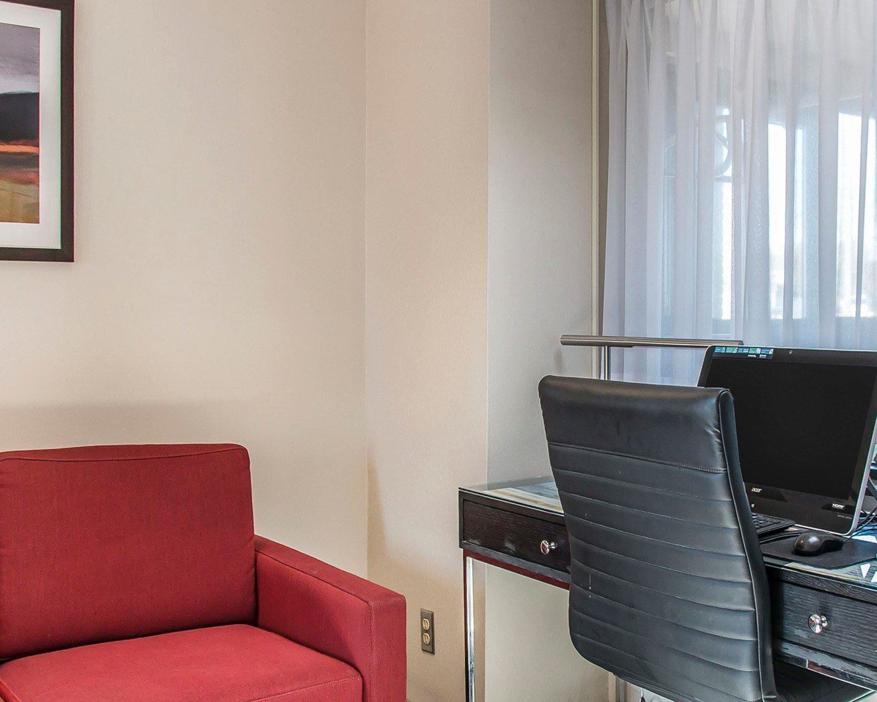 Habitación de hotel con un sillón rojo, una silla de oficina negra, un escritorio con una computadora portátil y una ventana con cortinas traslúcidas.