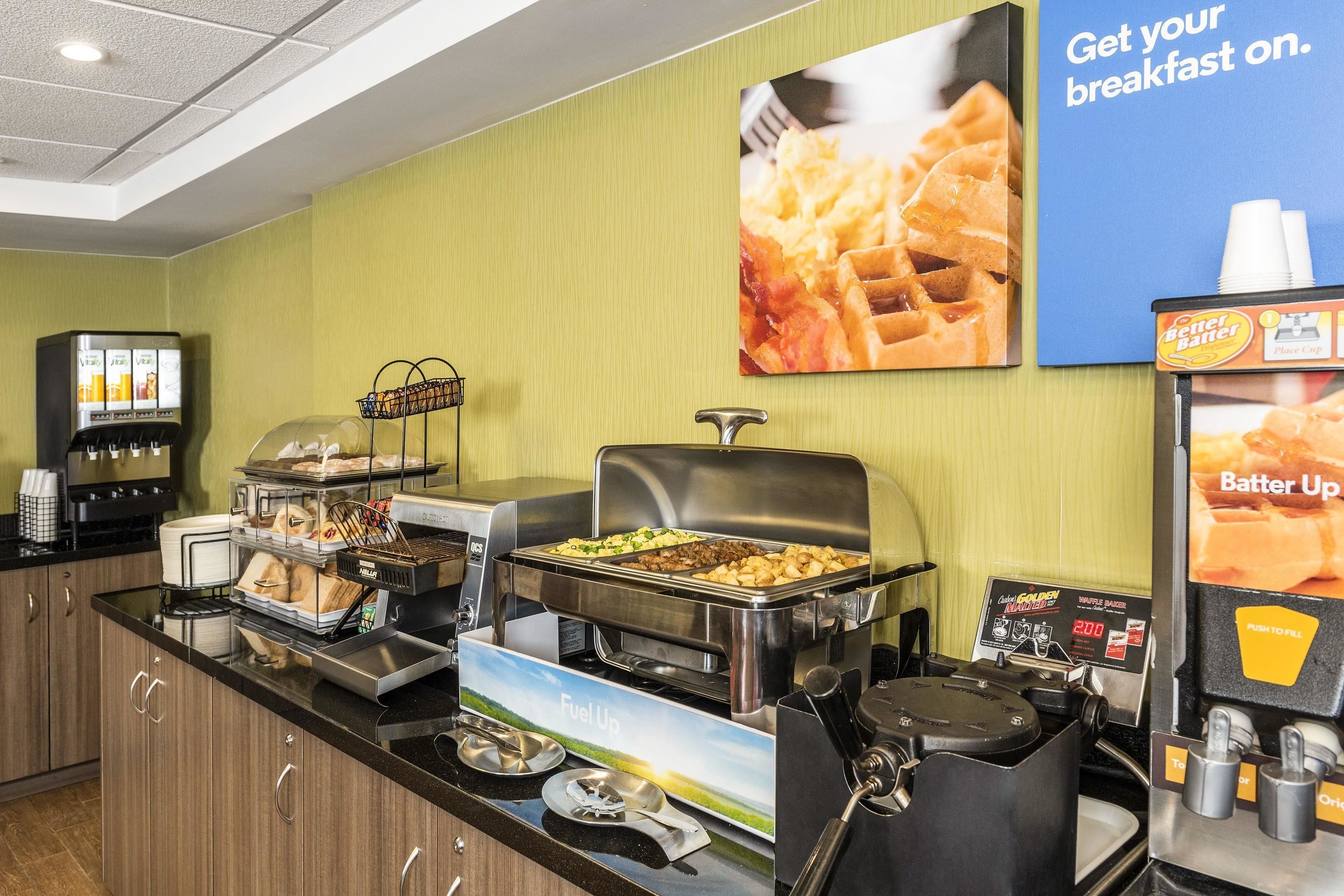 Montaje de bufé con diversos alimentos, una máquina de café y un cartel anunciando el desayuno.