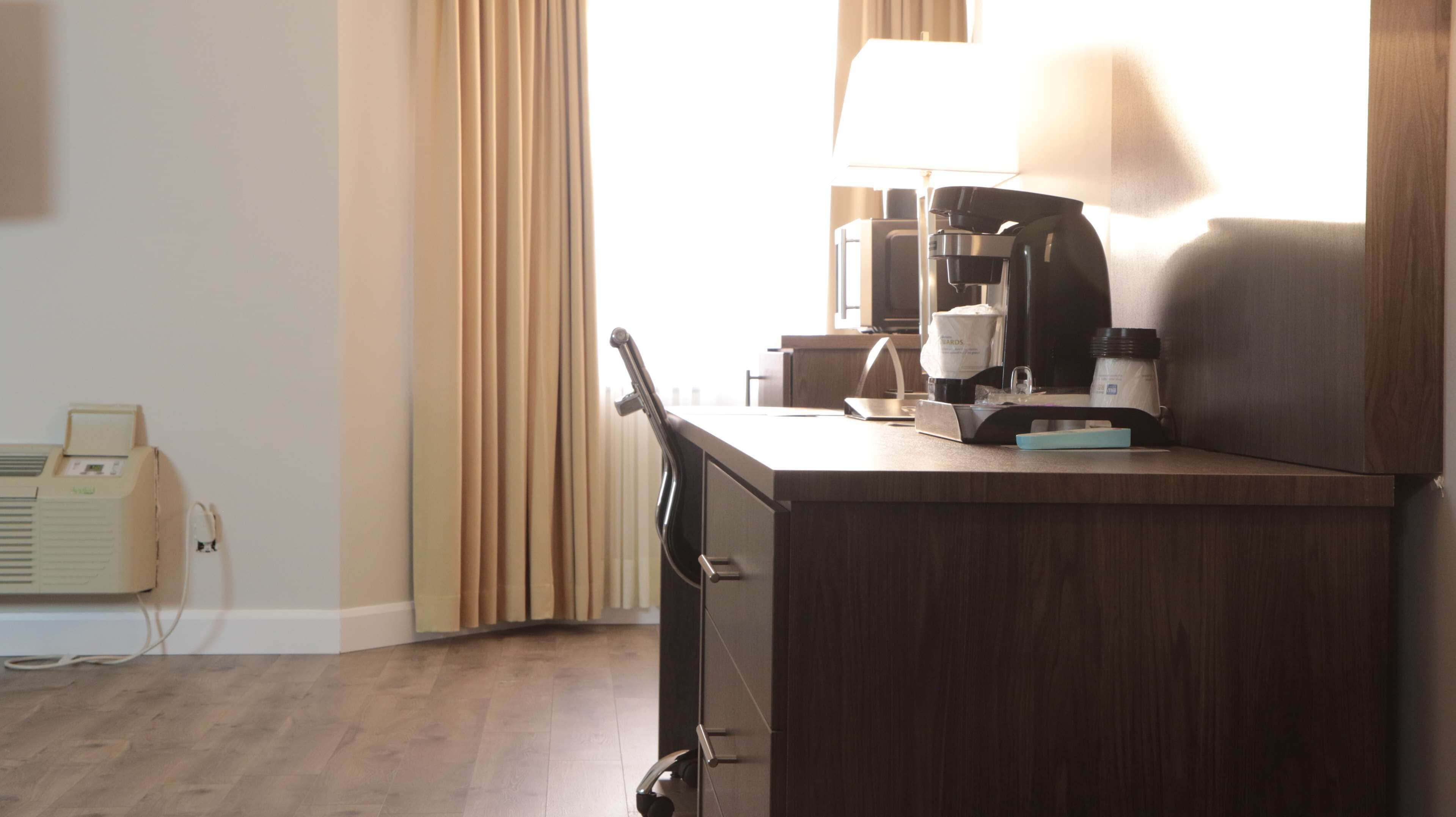 A hotel room with a desk, a coffee maker, a lamp, a telephone, and a window with curtains.
