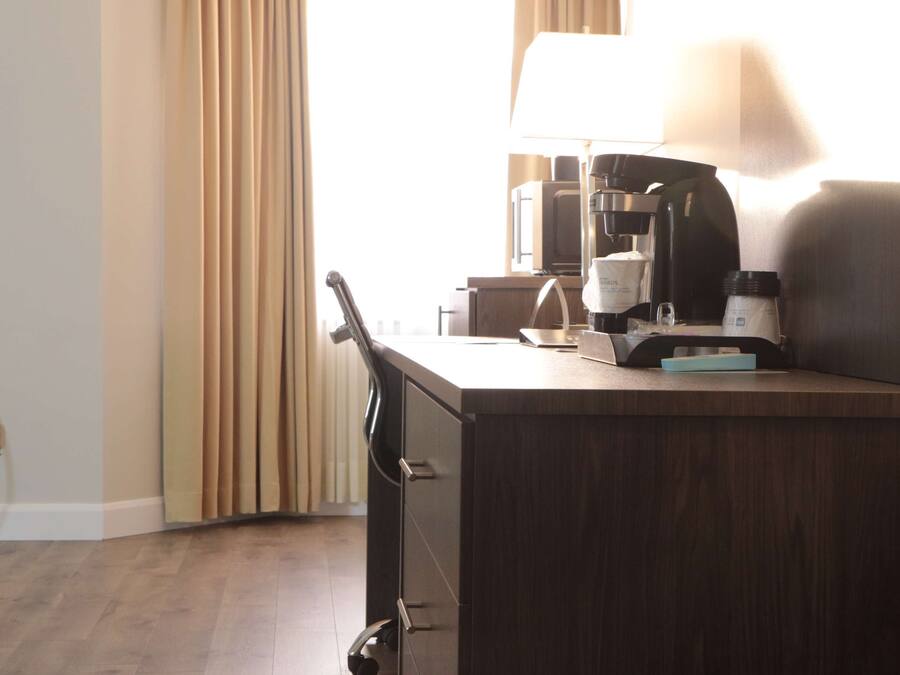 A hotel room with a desk, a coffee maker, a lamp, a telephone, and a window with curtains.