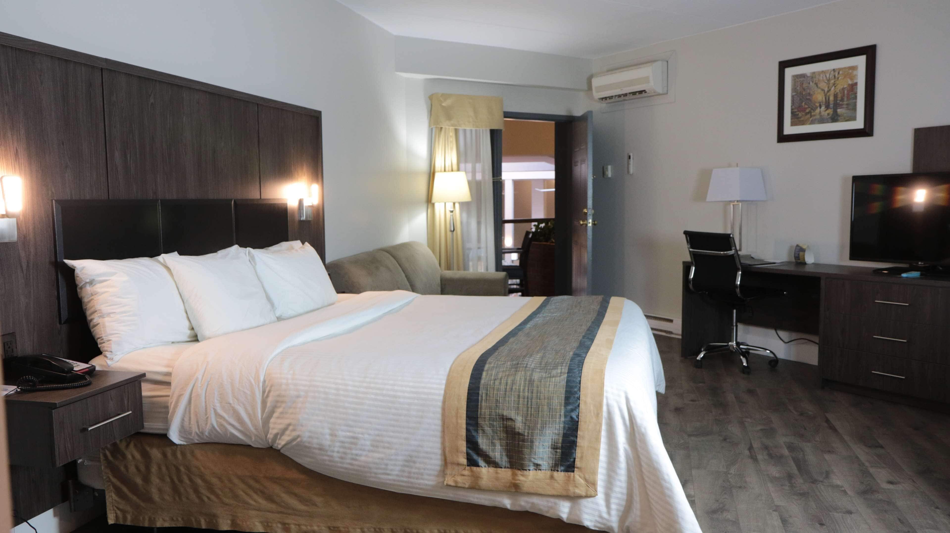 A hotel room with a bed, a desk, a chair, a TV, and an air conditioning unit.