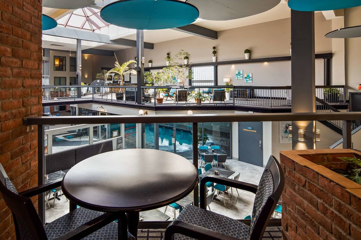 A modern indoor space with a glass railing, a round table, and chairs, overlooking a lower level with seating and plants.