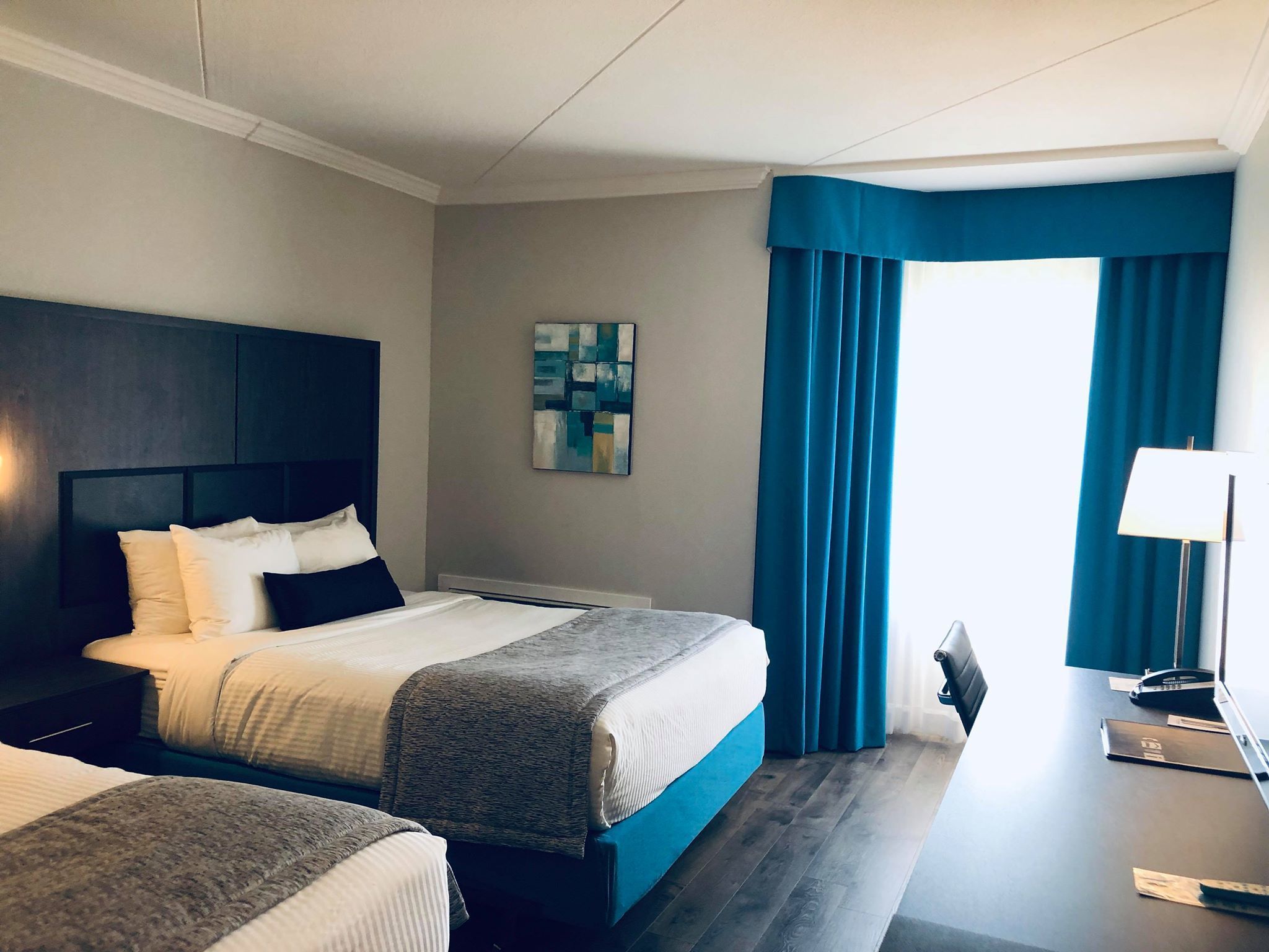 A hotel room with two beds, a desk with a lamp, a painting on the wall, and a large window with blue curtains.