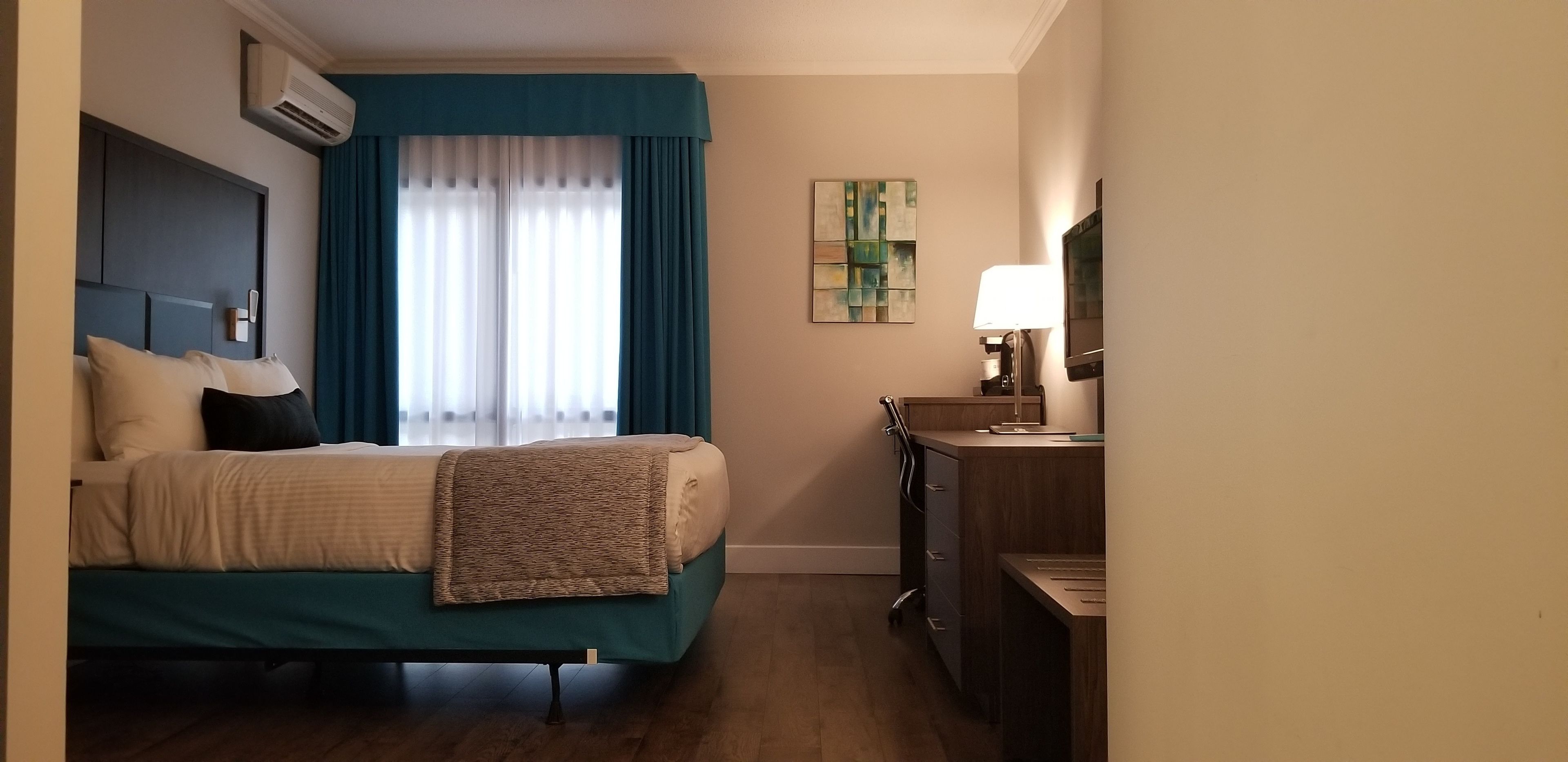 Une chambre d’hôtel avec un lit, un bureau, un climatiseur, une lampe et un tableau au mur.