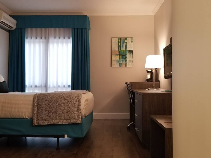 A hotel room with a bed, a desk, an air conditioner, a lamp, and a painting on the wall.
