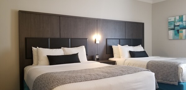 Habitación de hotel con dos camas, cabeceros de madera oscura, ropa de cama blanca y una lámpara fijada a la pared.