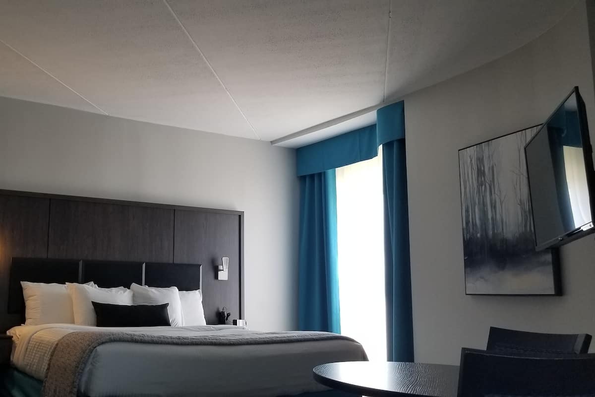 A modern hotel room with a large bed, a bedside table, a small round table, and a large window with curtains.