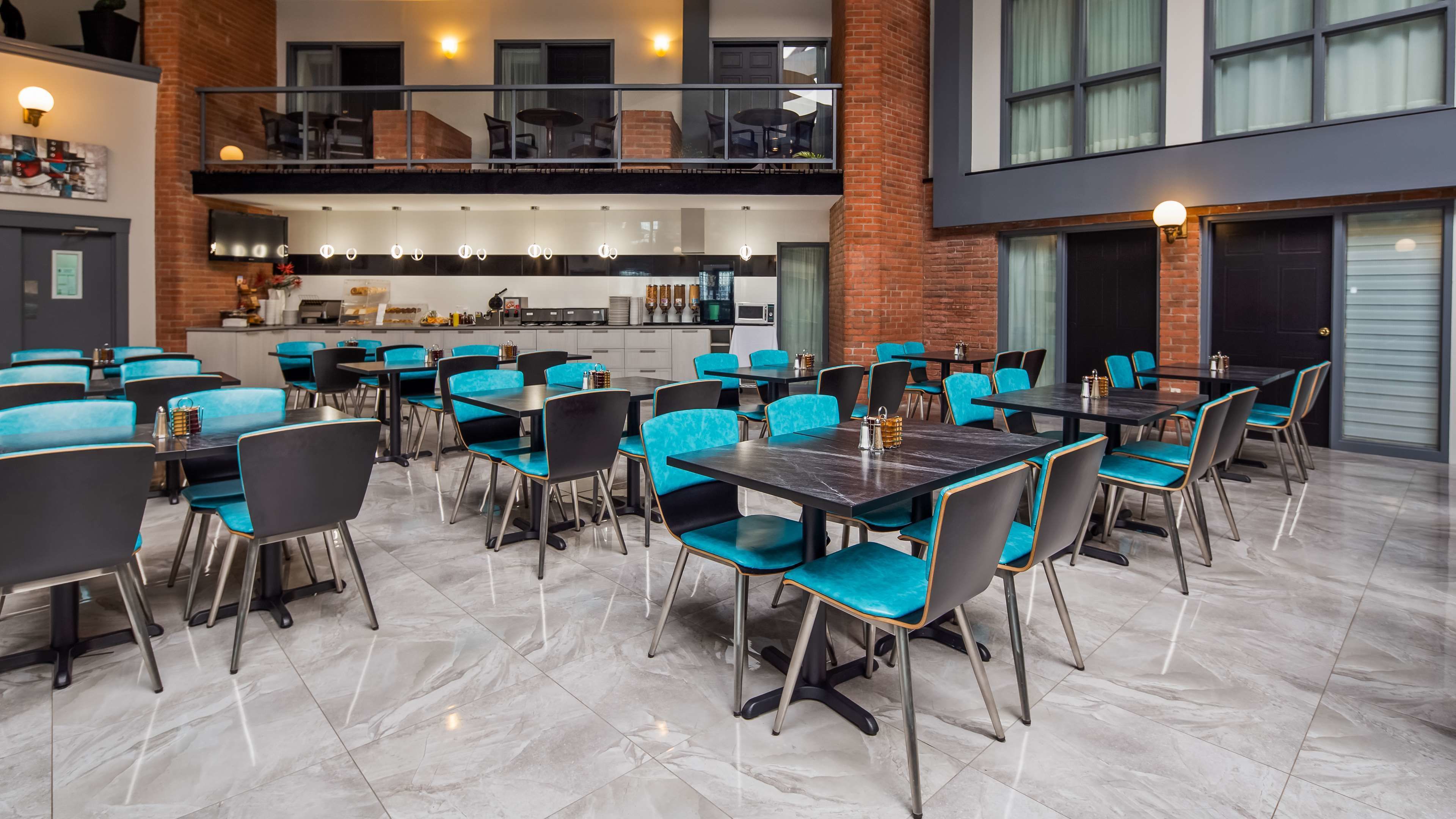 A modern dining area with multiple tables and chairs, a kitchen in the background, and a brick wall.