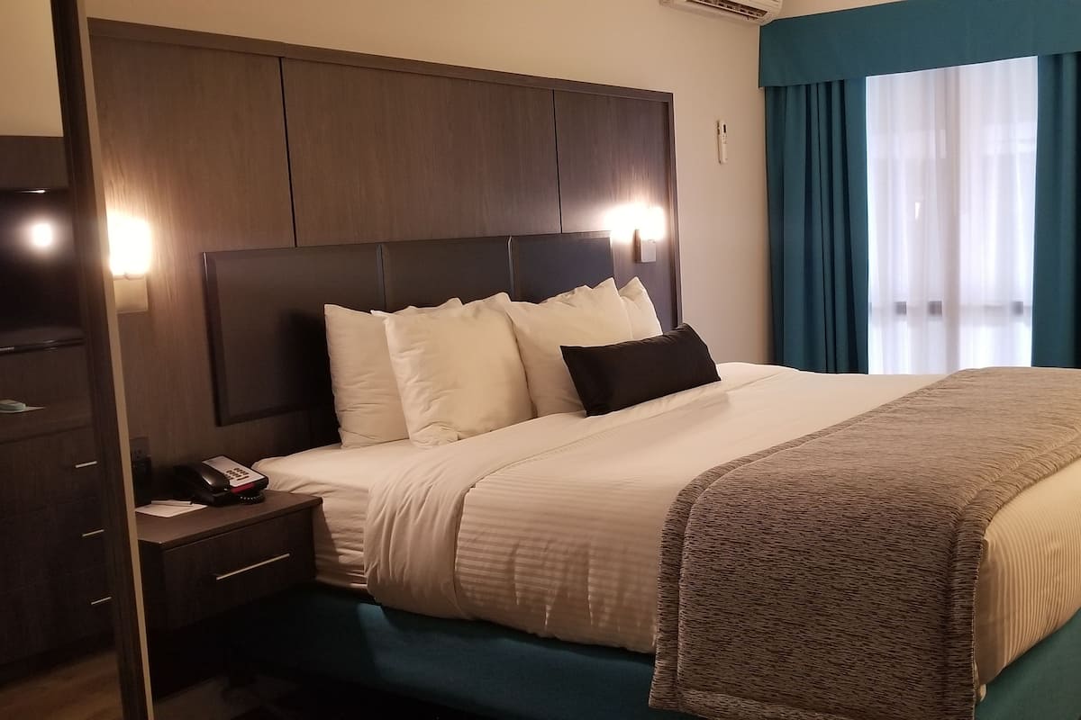 A hotel room with a large bed, a bedside table with a phone, a nightstand, a dark headboard, and a window with blue curtains.