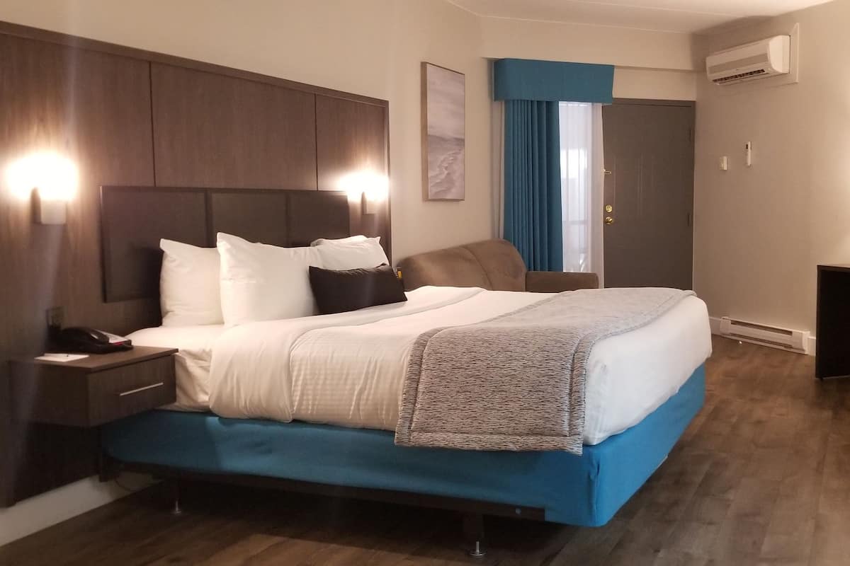 A hotel room with a large bed, a nightstand, a desk, and an air conditioning unit.