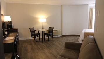A hotel room with a wooden floor, a bed, a sofa, a desk with a lamp, and a chair.