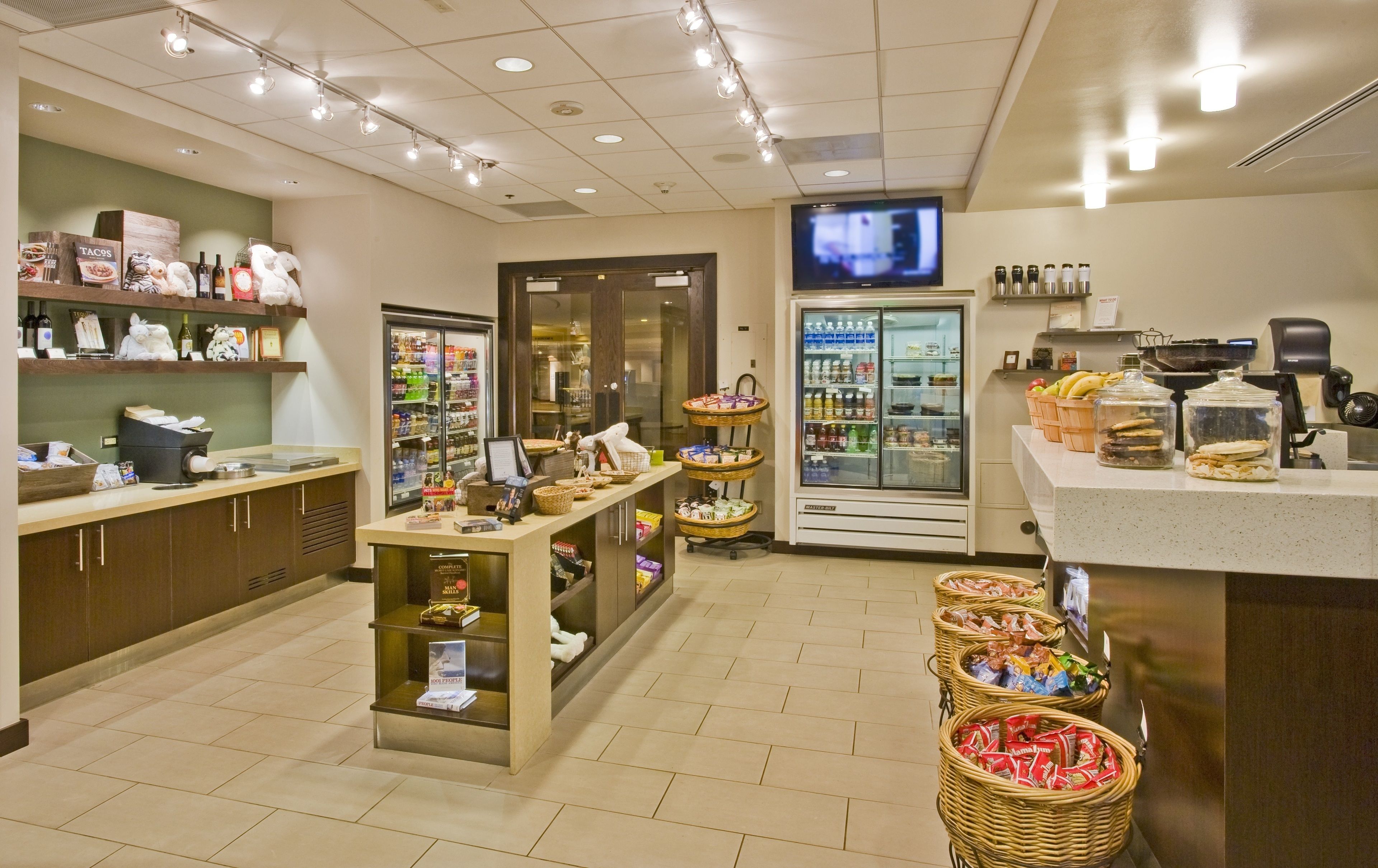 Una cafetería bien iluminada con mostrador, estanterías con aperitivos, un refrigerador y una pantalla de televisión.