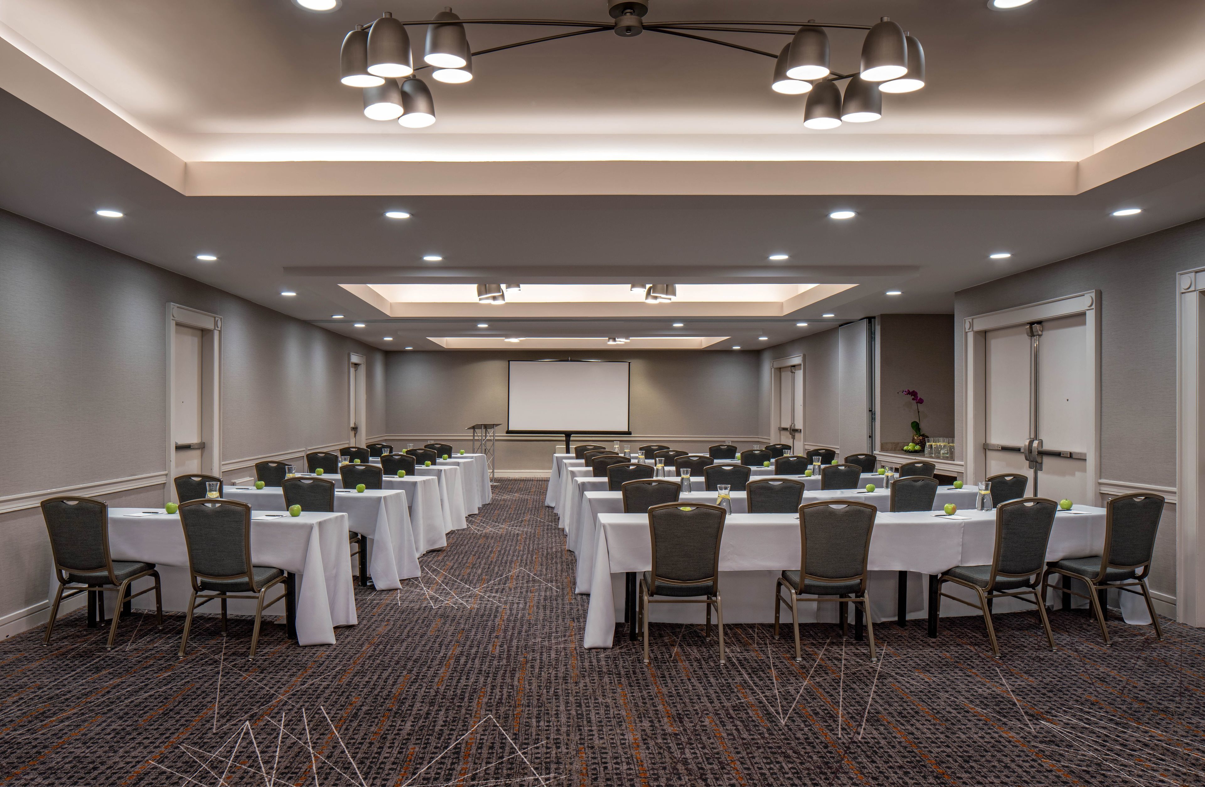 Sala de conferência com várias mesas e cadeiras, equipada com projetores e luminárias.