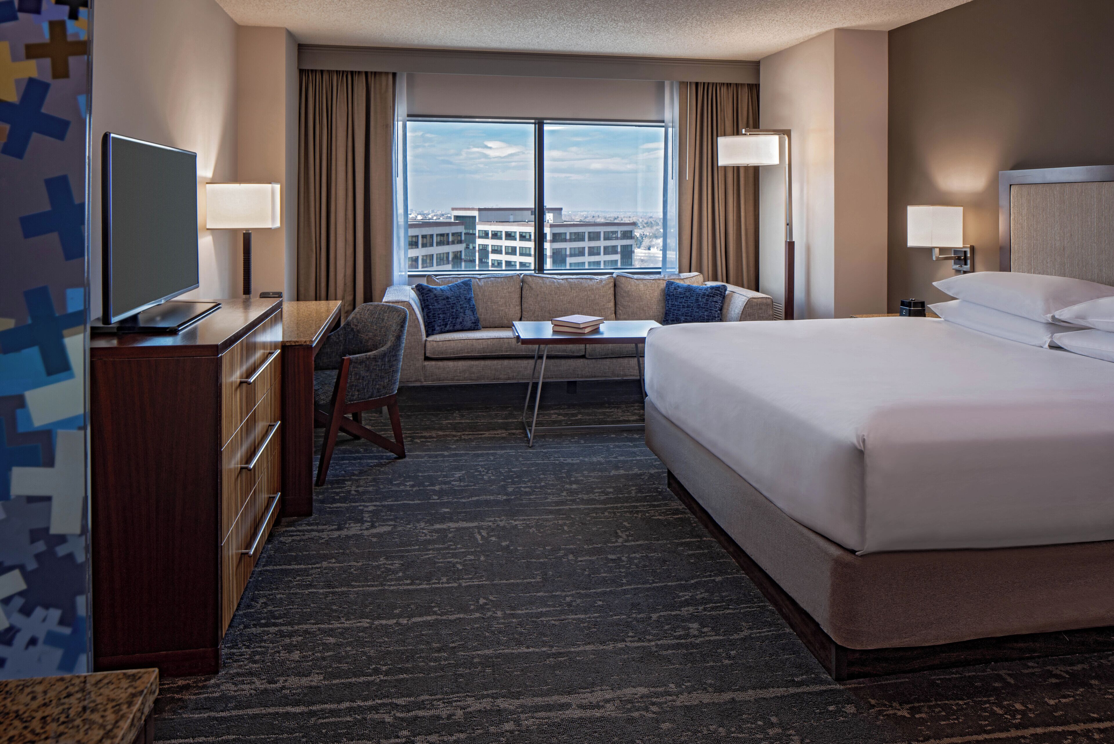 A hotel room with a large bed, a sofa, a desk with a TV, and a view of the city through large windows.