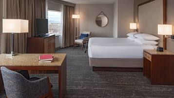 A hotel room with a bed, a desk with a chair, a television, and a view of the city through the window.