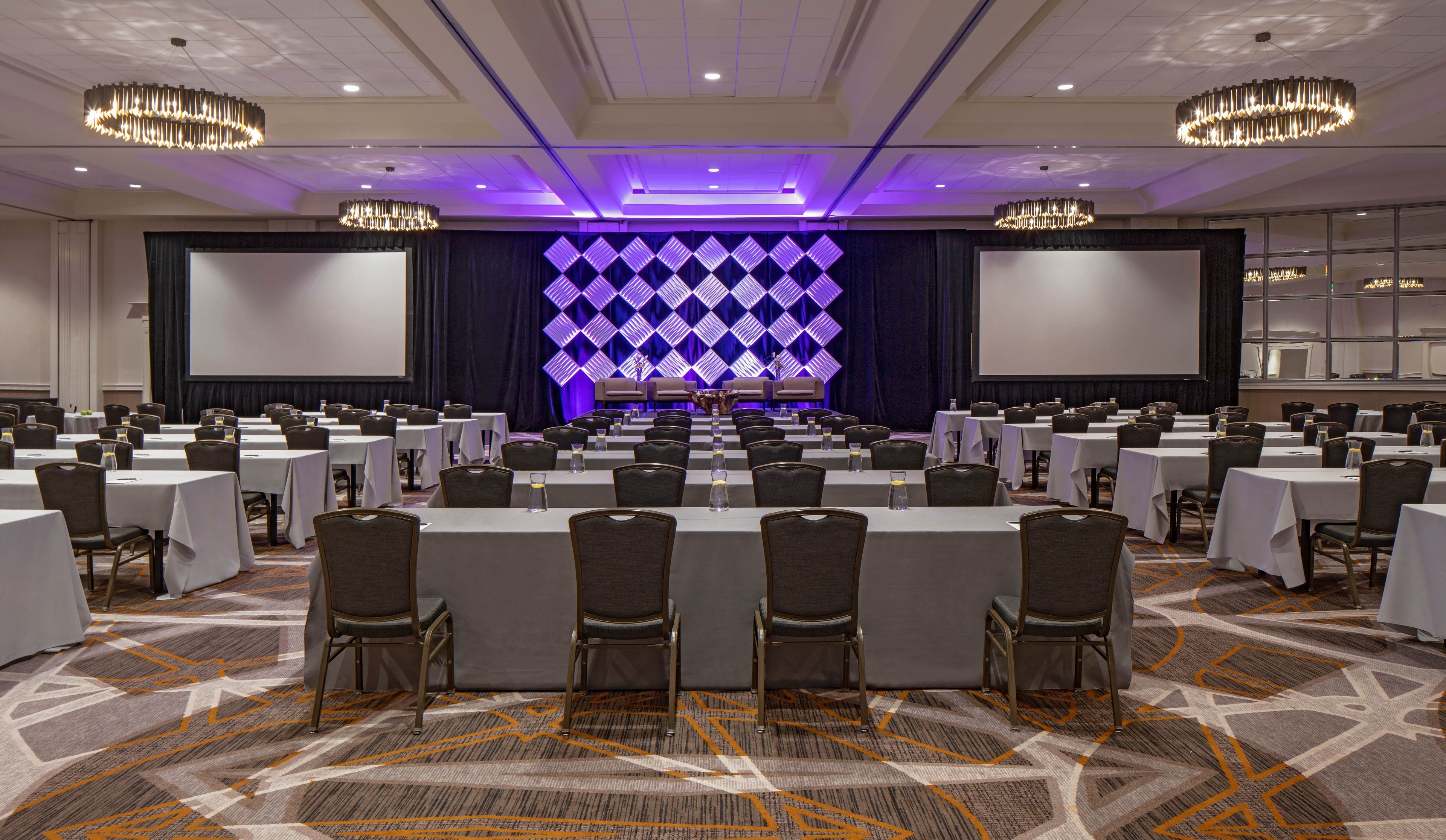 A conference room with rows of tables and chairs, a stage with a blue geometric backdrop, and chandeliers.