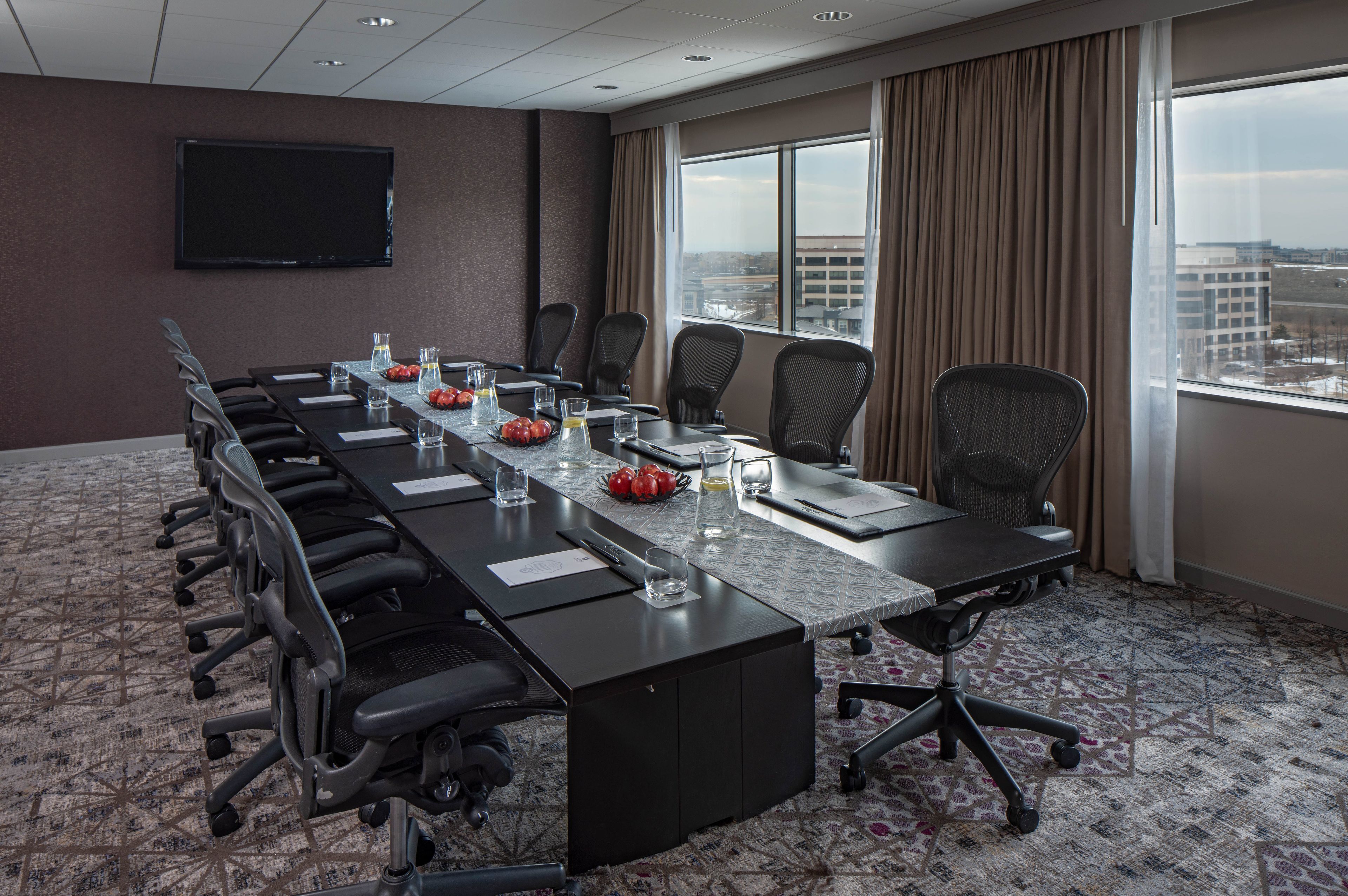 A conference room with a long table, chairs, a flat-screen TV, and a city view through large windows.