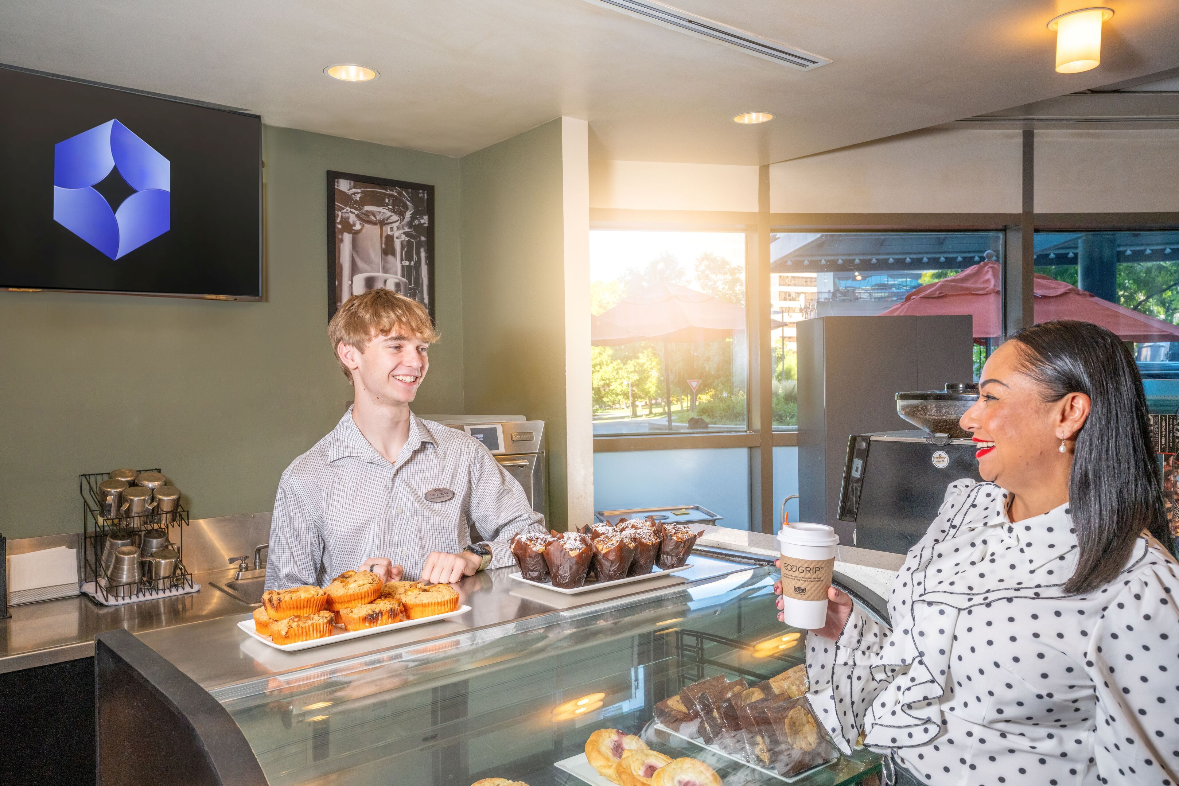 A bakery counter with pastries and a coffee cup, a large window with a view of the outside, and a logo on the wall.