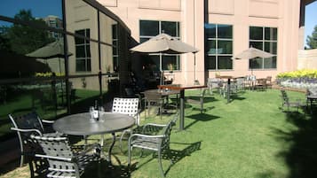 An outdoor patio area with tables and chairs, surrounded by a building with large windows and a well-maintained lawn.