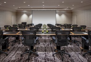Un salón de conferencias con una larga mesa, varias sillas, una pantalla de proyector y suelo alfombrado.