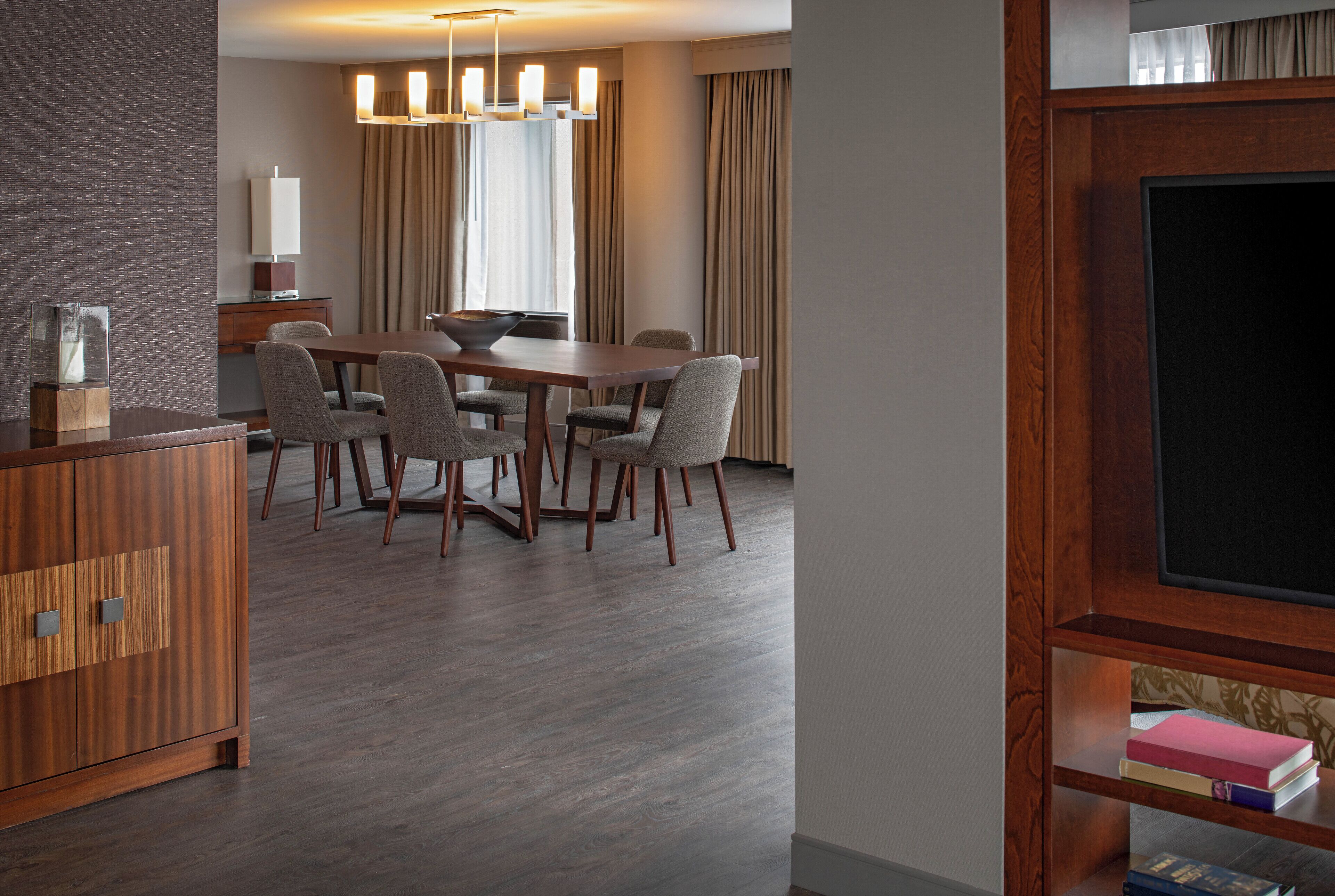 A modern hotel room with a wooden dining table, four chairs, a wooden cabinet, a flat-screen TV, and a bookshelf.