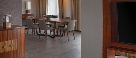 A modern hotel room with a wooden dining table, four chairs, a wooden cabinet, a flat-screen TV, and a bookshelf.