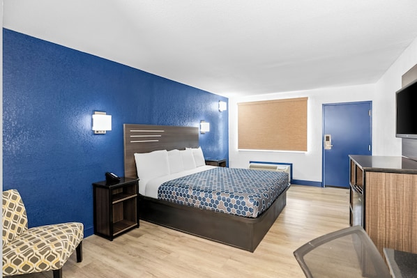 A hotel room with a bed, bedside table, chair, desk, TV, and a window with blinds.