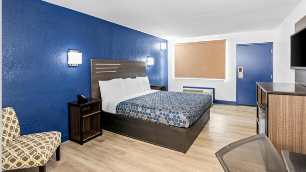A hotel room with a bed, bedside table, chair, desk, TV, and a window with blinds. - Americas Best Value Inn Richmond South (Richmond)