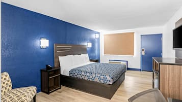A hotel room with a bed, bedside table, chair, desk, TV, and a window with blinds.