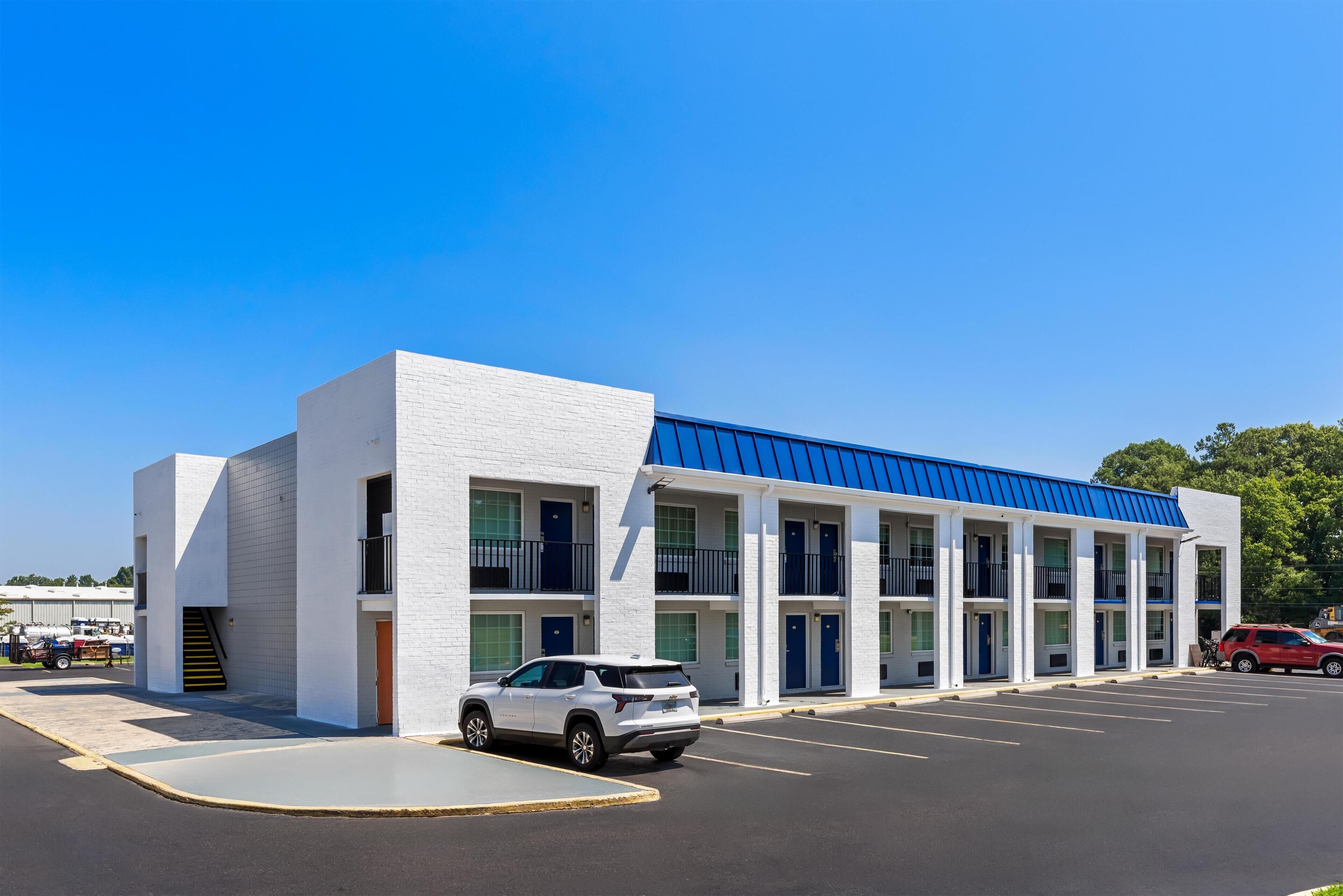 A multi-story hotel with a white facade and blue roof, featuring a parking lot with several vehicles.