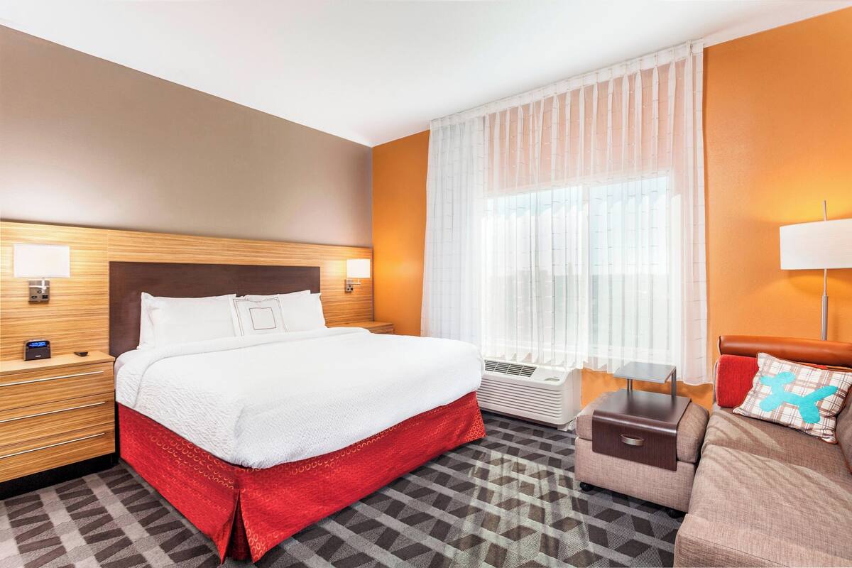 Una habitación de hotel con una cama grande, una mesita de noche con lámpara, un sofá pequeño y una ventana con cortinas translúcidas.