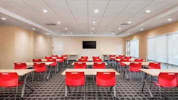 Un salón de conferencias con varias mesas y sillas rojas, un televisor de pantalla plana en la pared y ventanas amplias en un lateral.