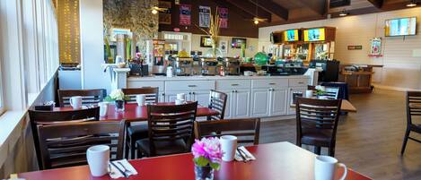 A restaurant with multiple dining tables and chairs, a bar area, and a kitchen in the background.