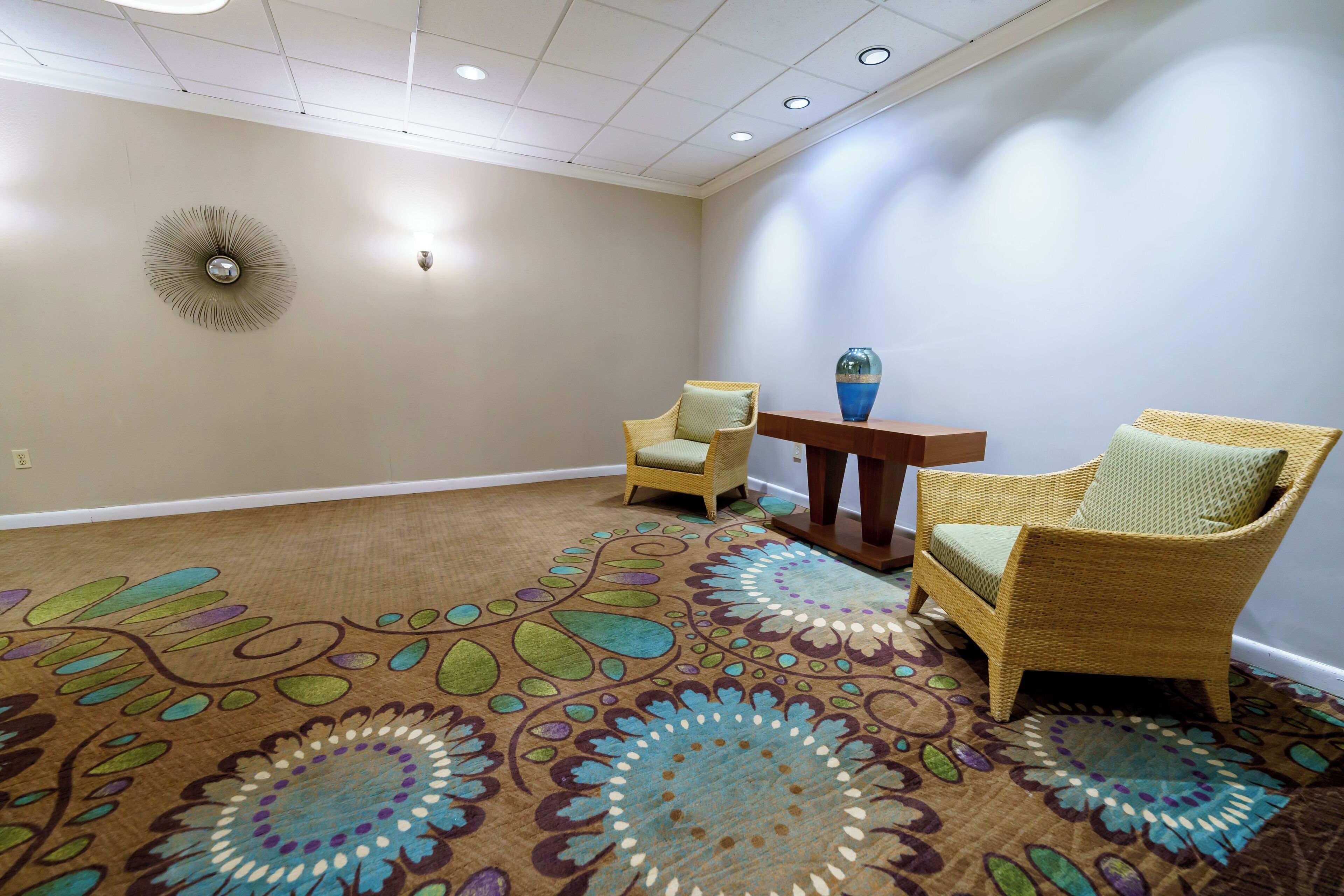 Une pièce recouverte de moquette, avec deux fauteuils en osier et une petite table, un élément décoratif mural et des spots encastrés au plafond.