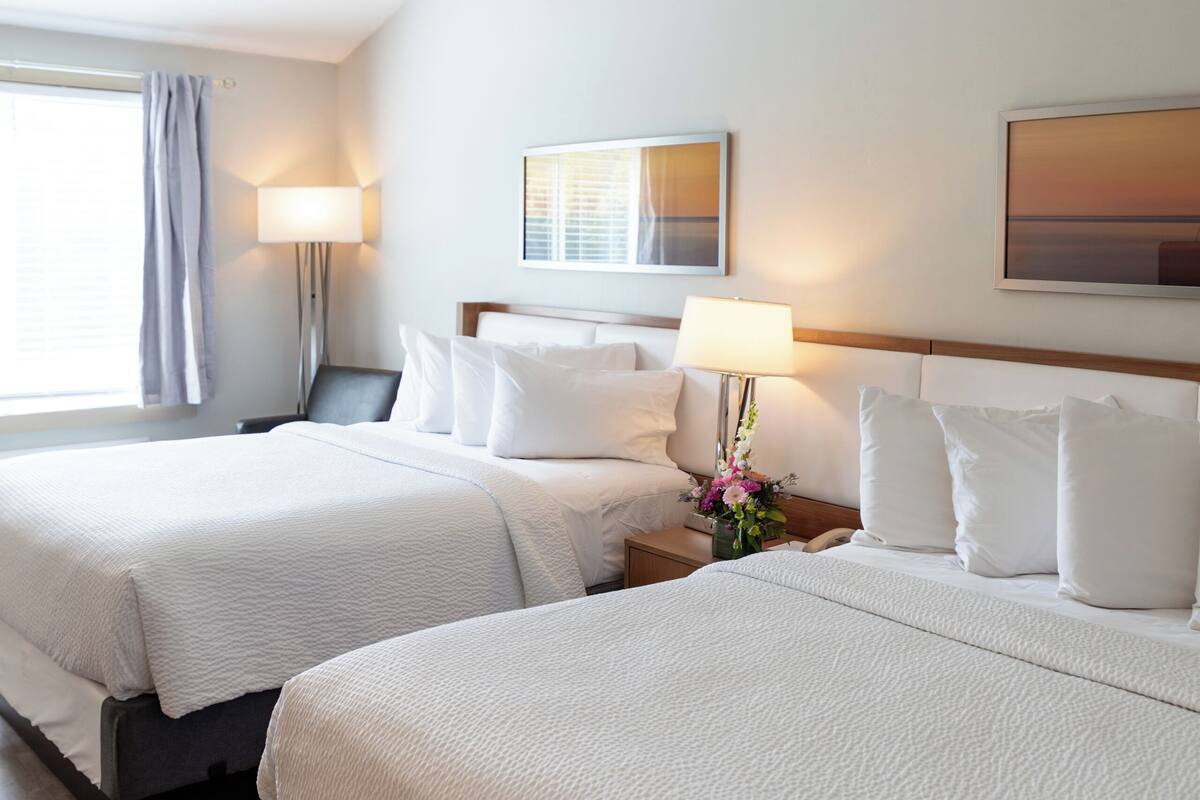 A hotel room with two beds, a nightstand with a lamp, a wall-mounted artwork, and a window with curtains.