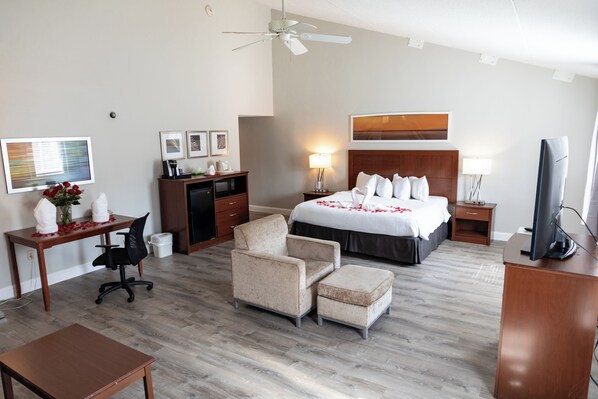 A hotel room with a bed, desk, chair, ottoman, TV, and ceiling fan.