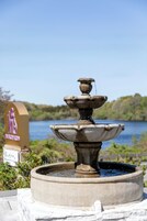 A stone fountain with water cascading from its tiers, set against a backdrop of a body of water and greenery.