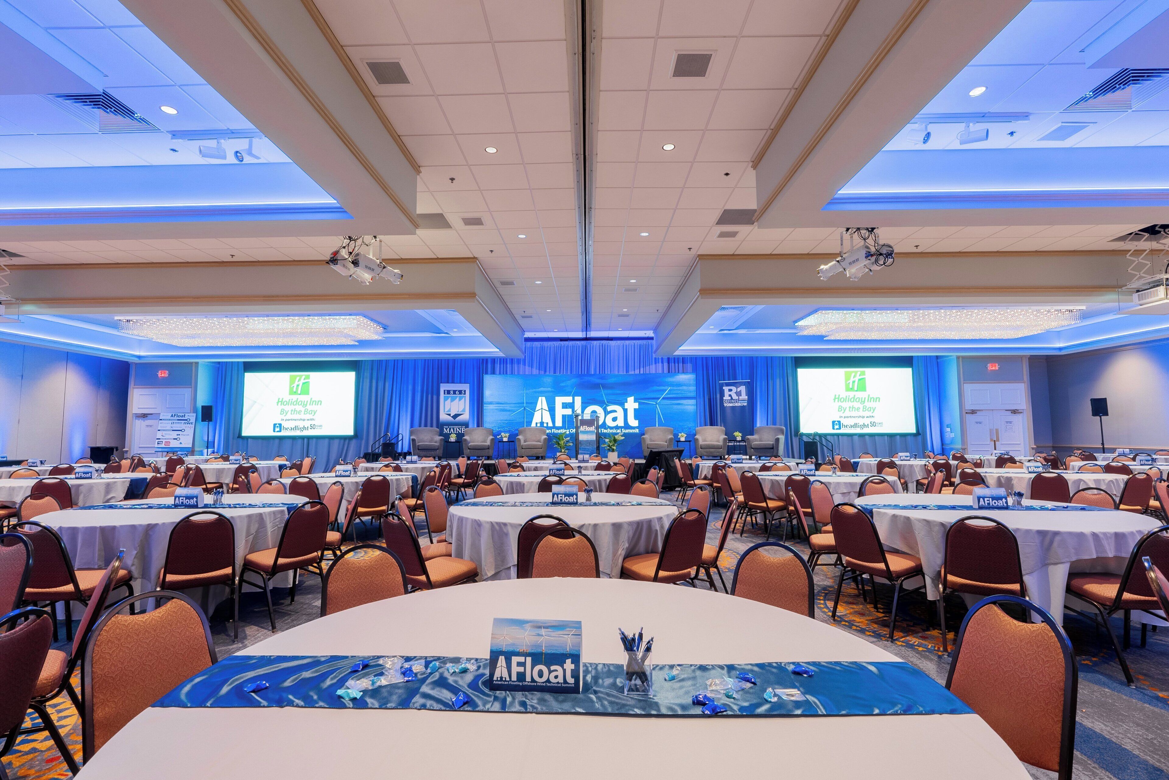 A conference room with multiple tables and chairs, a stage with a presentation screen, and a banner reading 'AFloat American Floating Offshore Wind Technical Summit'.