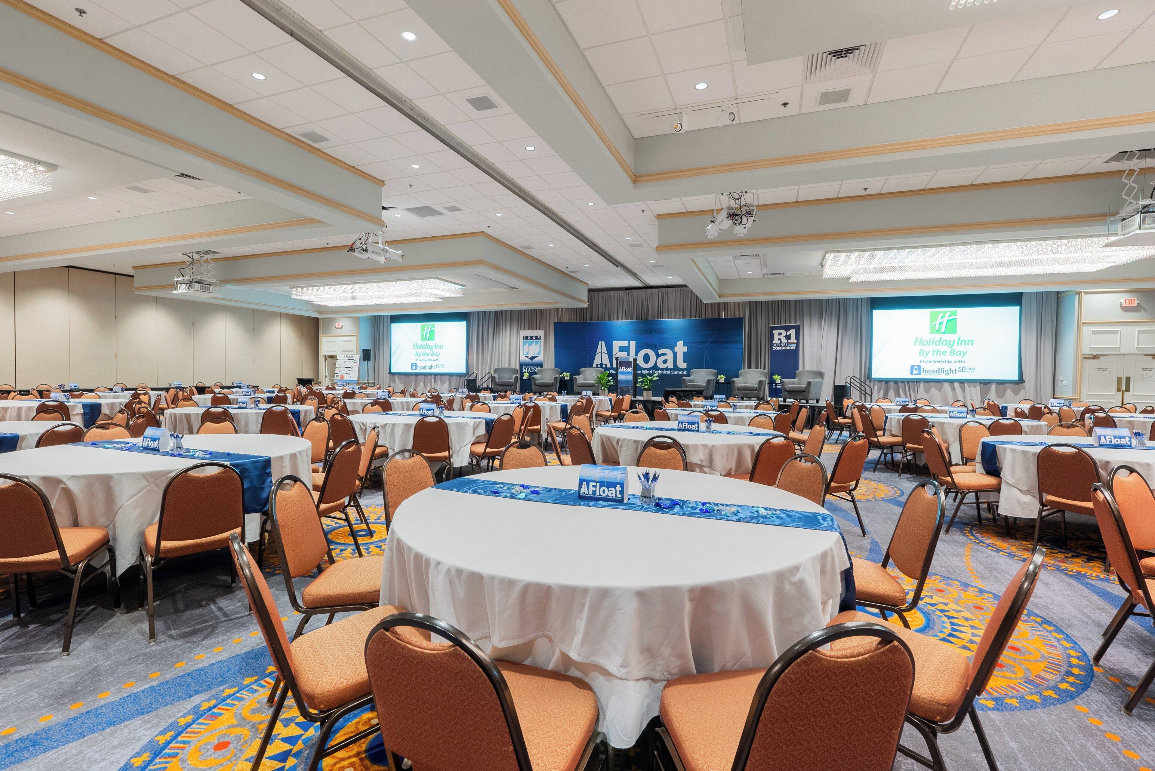 A large conference room with round tables and orange chairs, featuring a blue and white color scheme and a carpeted floor.
