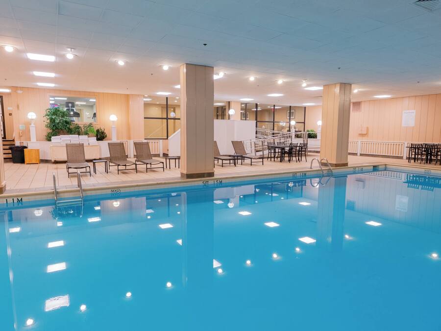 Una piscina con sillas de descanso y mesas, rodeada por un espacio interior bien iluminado y con vistas al exterior.