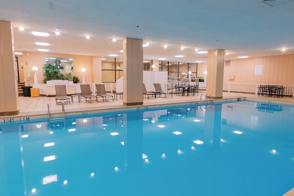 Piscina com espreguiçadeiras e mesas, rodeada por um espaço interior bem iluminado e com vista para o exterior.