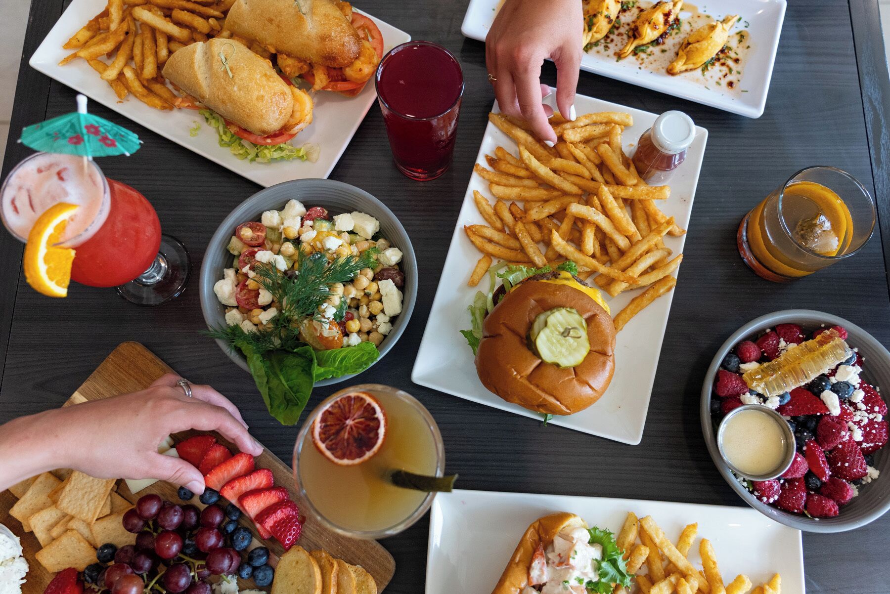 Une variété d’aliments, notamment des sandwichs, des frites, une salade et une assiette de fruits, sont disposés sur une table.