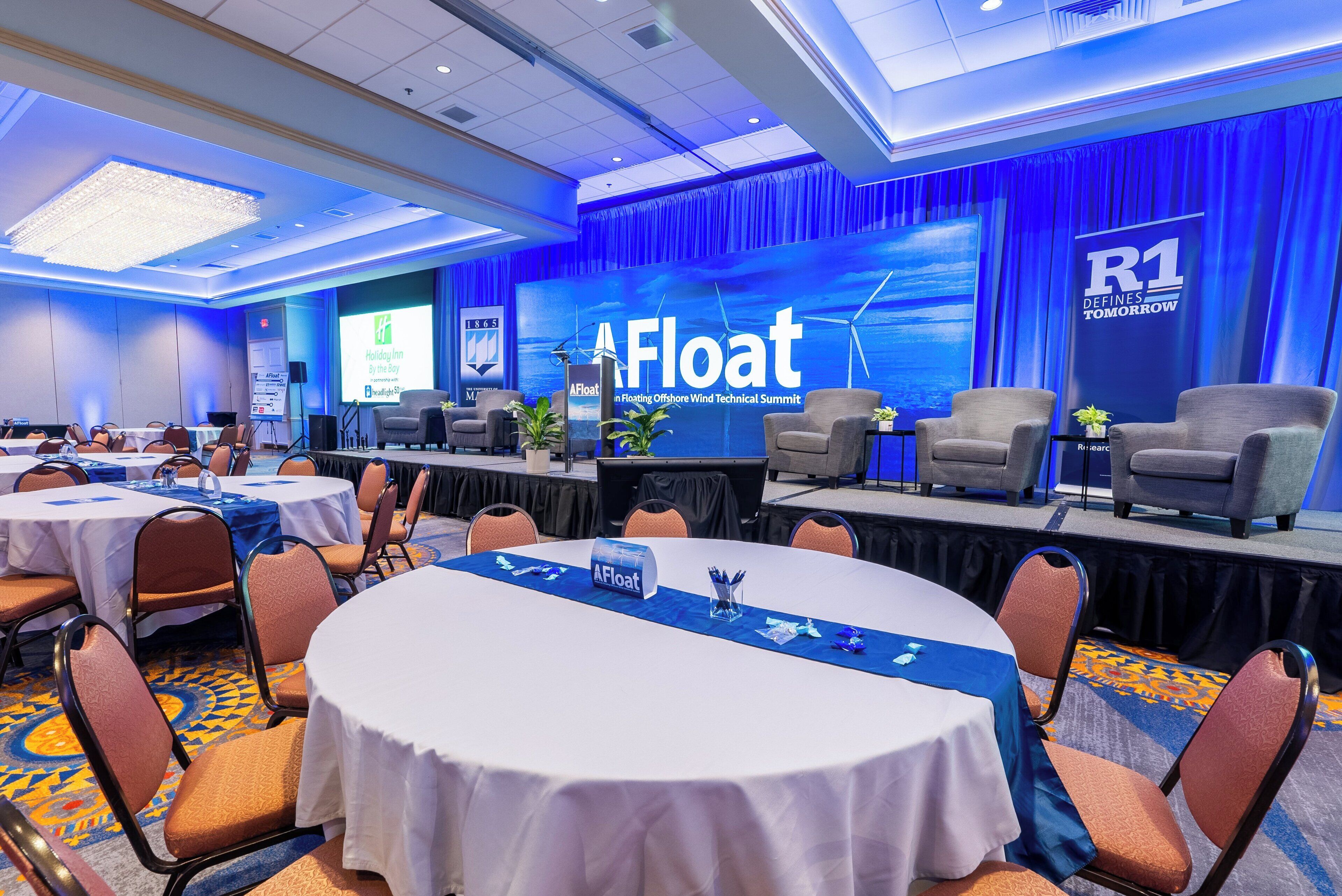 A conference room with round tables, chairs, and a stage with a presentation screen.