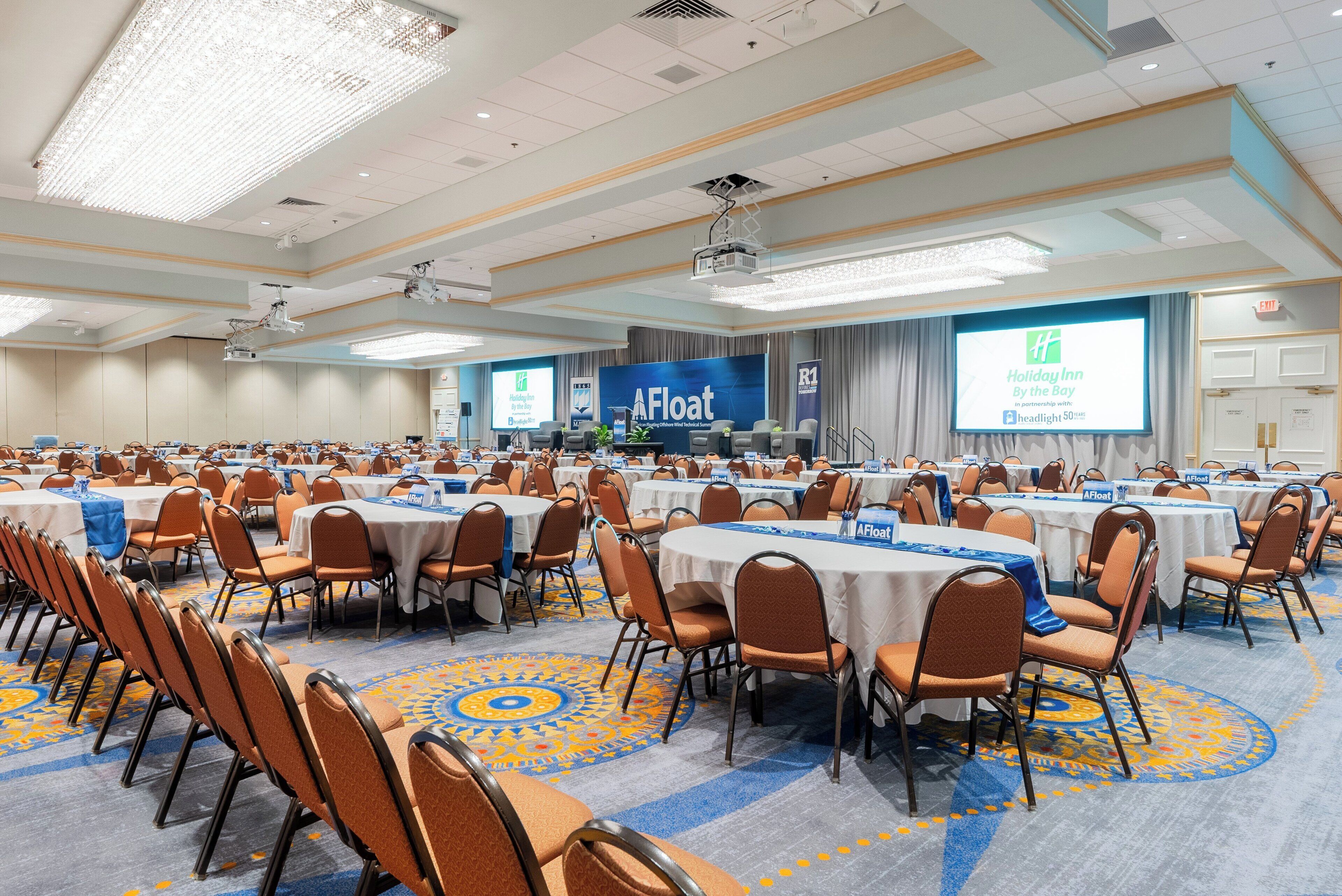 A conference room with multiple tables and chairs, a large screen displaying a presentation, and a stage with a podium.