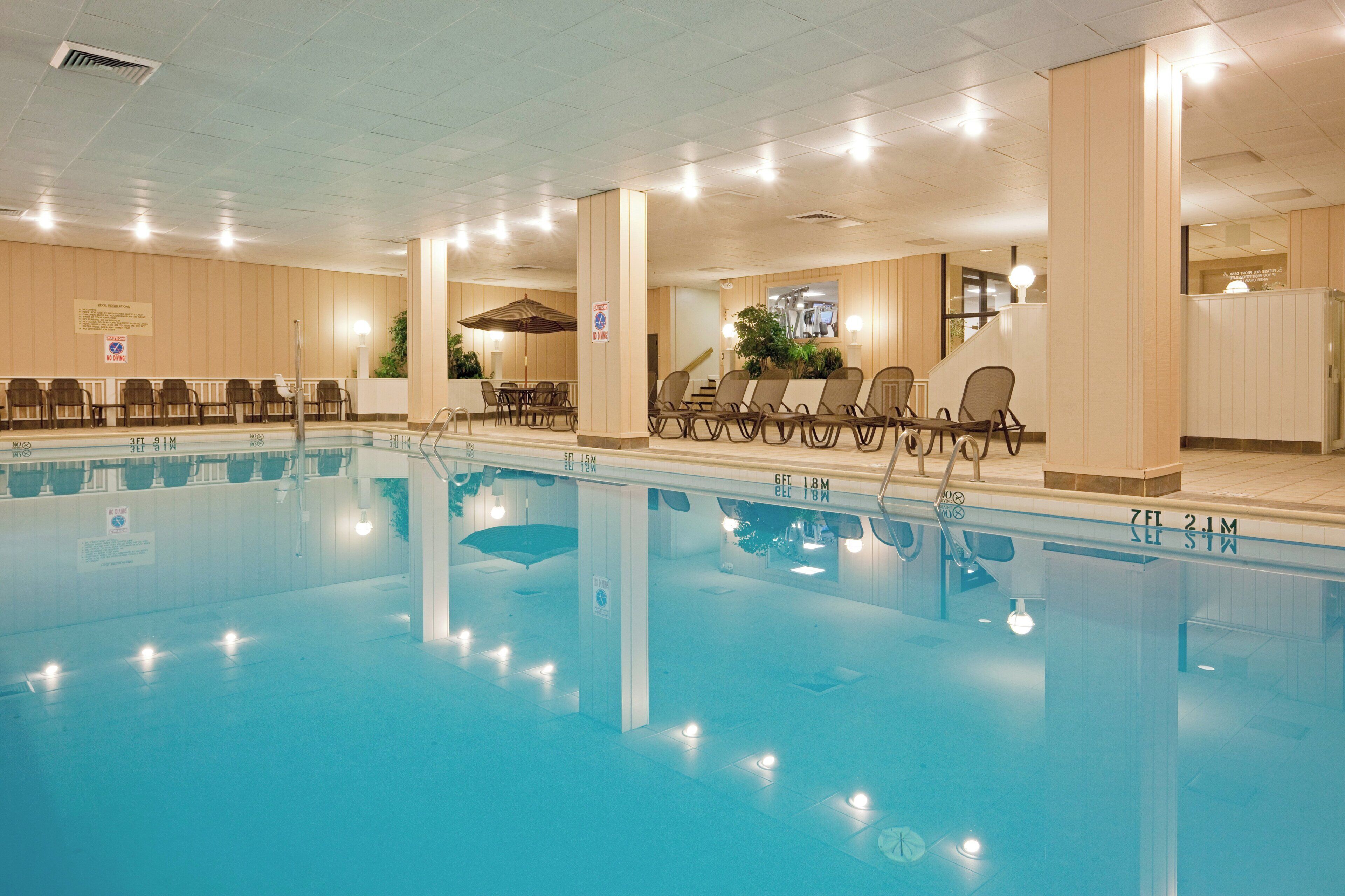 A swimming pool with lounge chairs and tables, indoor lighting, and a ceiling with ventilation.