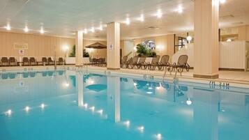 A swimming pool with lounge chairs and tables, indoor lighting, and a ceiling with ventilation.