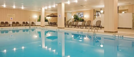 Una piscina con sillones de descanso y mesas, iluminación interior y un techo con ventilación.