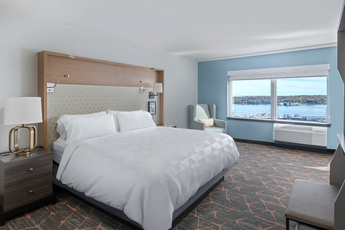 A hotel room with a large bed, a nightstand, a desk, a chair, and a window with a view of a body of water.