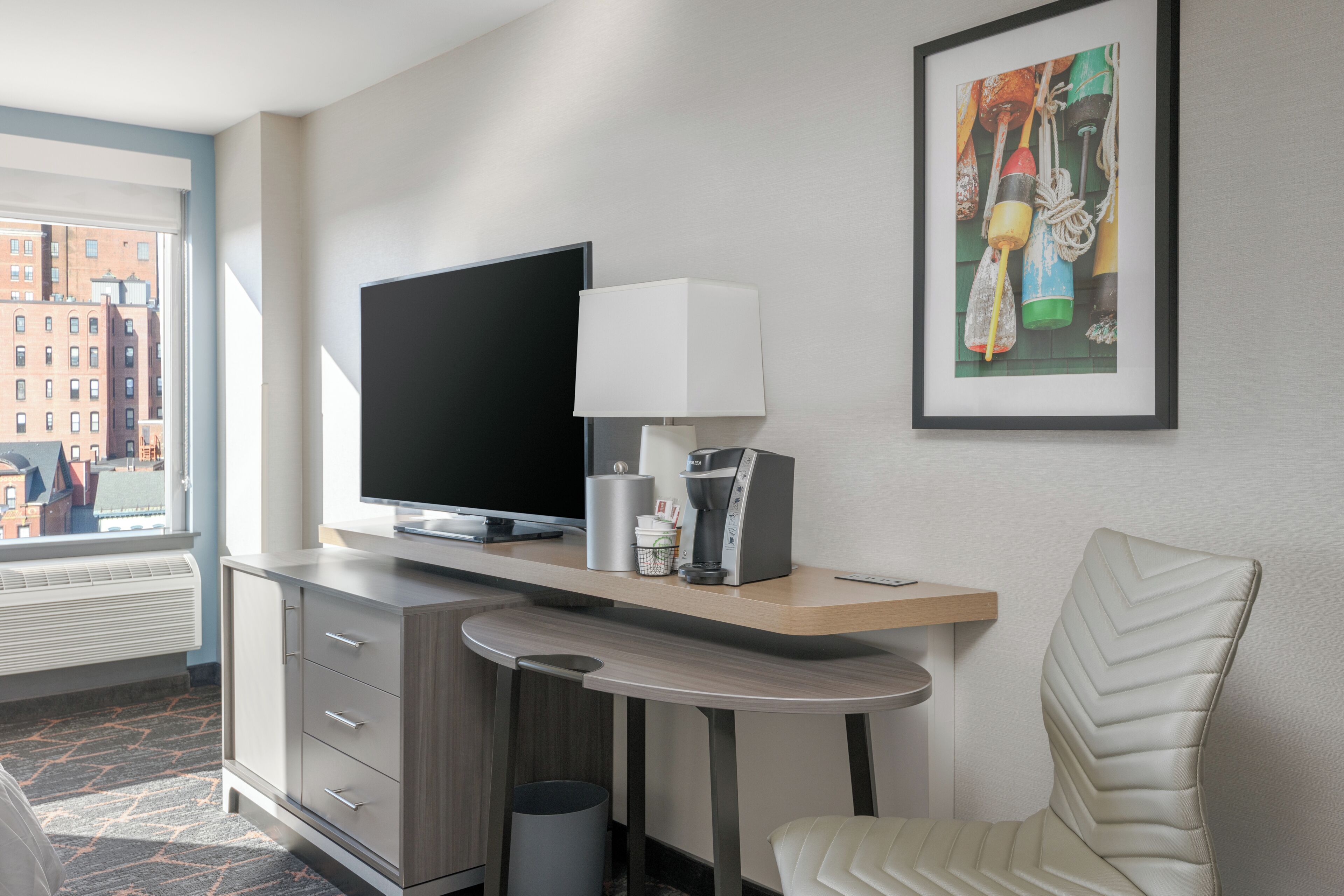 A hotel room with a flat-screen TV, a coffee maker, a framed artwork, a chair, and a desk.