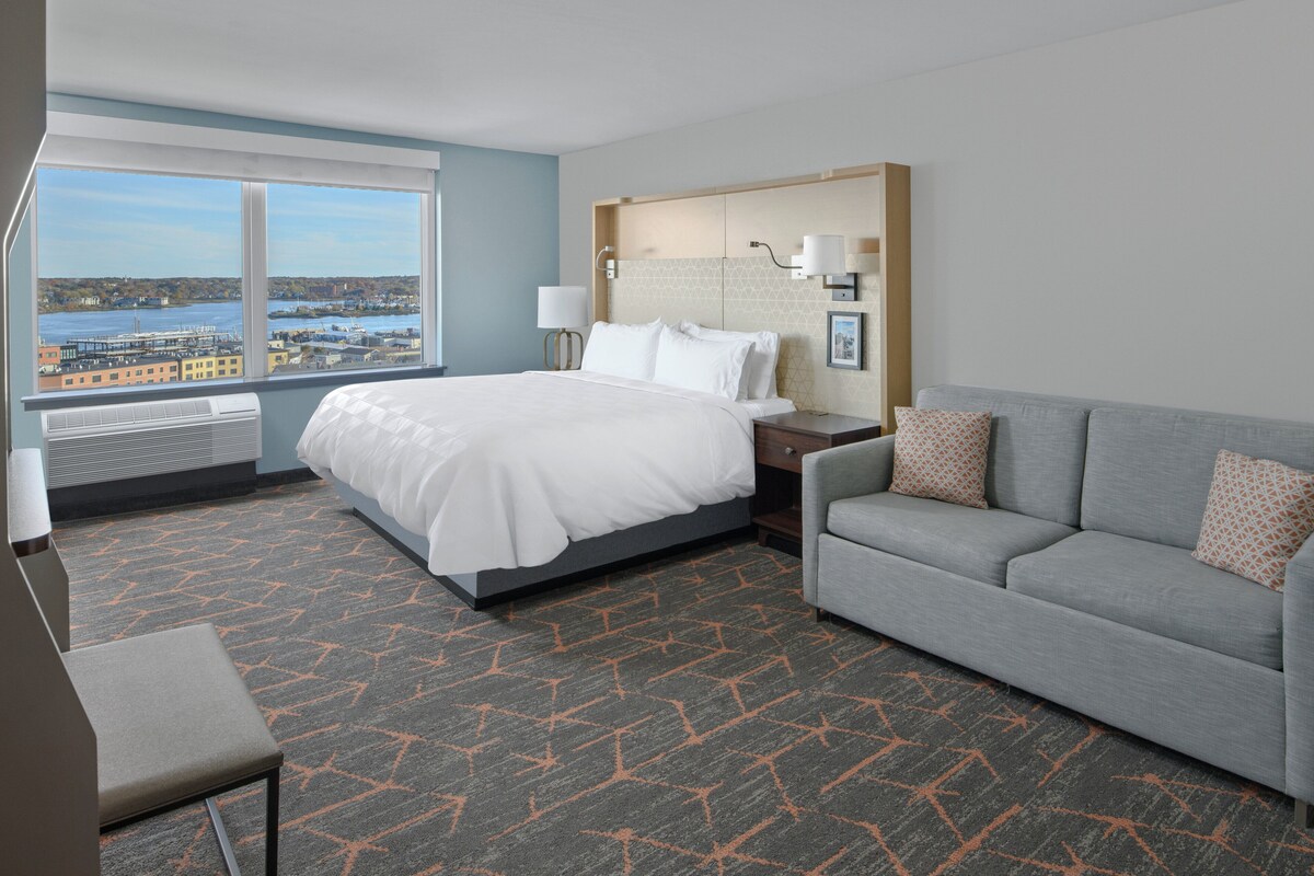 A hotel room with a large bed, a grey sofa, and a view of a cityscape through large windows.